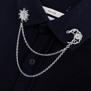 May include: Silver-toned collar chain with sun and moon charms. The sun charm is on the left, and the moon charm is on the right. Both charms have a small, round, light-colored stone in the center. The chain is attached to a black collared shirt.