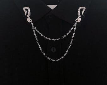 Könnte beinhalten: Silberfarbene Kragenclips mit Kettenanhänger, gezeigt auf einem schwarzen Hemd mit Kragen. Die Clips haben ein dekoratives, stilisiertes Design. Die Kette hängt zwischen den Clips und erzeugt einen geschichteten Effekt.