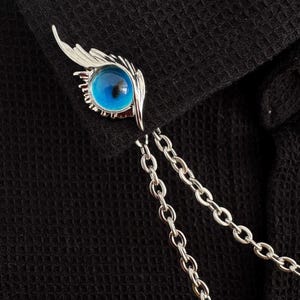 May include: A silver-toned lapel pin featuring a blue eye design with a feather accent. The pin has a chain that hangs down. The pin is set against a black fabric background.