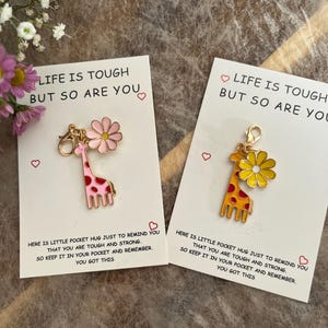 May include: Two giraffe charms with pink and yellow flower pendants on cards. The cards read "LIFE IS TOUGH BUT SO ARE YOU" and include a message about being tough and strong. The giraffes have red spots.