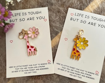 Op de afbeelding: Twee girafbedels met roze en gele bloemen aan kaarten. De kaarten lezen "HET LEVEN IS ZWAAR MAAR JIJ OOK" en bevatten een boodschap over sterk zijn. De giraffen hebben rode vlekken.