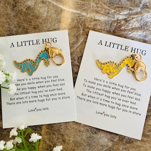 May include: Two cards with the text "A Little Hug" and a message. Each card has a dinosaur charm with a gold clasp. One dinosaur is blue and the other is yellow. White flowers are in the foreground.