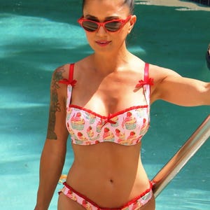 May include: Pink cupcake-print bikini set with red trim and bows. The top has adjustable straps and the bottoms have a ruffled edge. The swimwear is worn by a person with red sunglasses and a bun hairstyle.