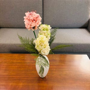 May include: A white ceramic vase with a pink and white flower arrangement. The vase is sitting on a wooden table.