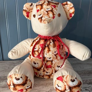 May include: A handmade teddy bear with a cream-coloured body and limbs. The head, ears, and parts of the paws and chest are patterned with repeating images of teddy bears and red hearts. A red polka-dot ribbon is tied around the neck.