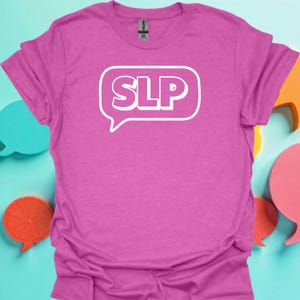 May include: A magenta t-shirt with the letters "SLP" inside a white speech bubble. The shirt is made of a soft material and has short sleeves. The shirt is displayed on a light blue background with colorful speech bubbles.