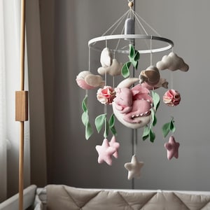 May include: A pink and white baby mobile with a crescent moon, stars, clouds, and leaves. A pink dragon is sleeping on the moon. The mobile is hanging from a wooden stand.
