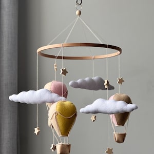 May include: A hanging mobile featuring three felt hot air balloons in mustard yellow, soft pink, and tan, suspended from a circular wooden hoop. The mobile includes white felt clouds and small wooden stars hanging on thin white strings from the frame.