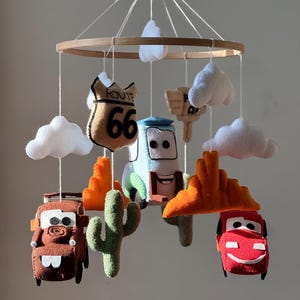 May include: A handmade baby mobile featuring felt car characters, clouds, cacti, and Route 66 signage. The mobile is suspended from a wooden ring with white string. The cars are various colors, including red, brown, and blue.