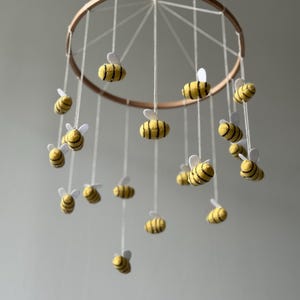 May include: A hanging mobile featuring multiple felt bees with yellow and black striped bodies and white wings, suspended by white strings from a circular wooden hoop. The bees are arranged at varying heights to create a layered effect.