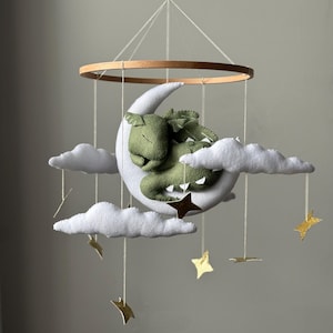 May include: A baby mobile featuring a green dragon sleeping on a white crescent moon. The mobile includes white felt clouds and gold star accents, suspended from a wooden ring.