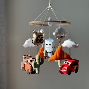 May include: A baby mobile featuring felt characters from the movie Cars, including Mater, Lightning McQueen, and others. The mobile includes Route 66 signage, clouds, and desert landscape elements. The mobile is suspended from a wooden ring.