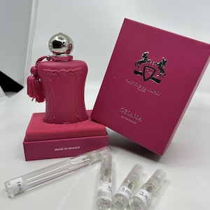 May include: A pink bottle of perfume with a silver cap and a tassel. The bottle is sitting on a pink box that says "Made in France". The box is next to a pink box with a silver crest and the words "The Different Company" and "Oriana Eau de Parfum". There are four small clear glass bottles with labels that say "10ml", "5ml", "3ml", and "2ml".