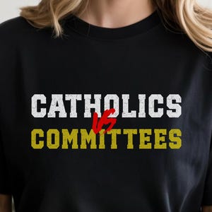 Op de afbeelding: Zwart t-shirt met de tekst "CATHOLICS vs COMMITTEES" in witte en gele letters. Het woord "vs" is rood. Het shirt is een casual stijl, geschikt voor dagelijks gebruik.