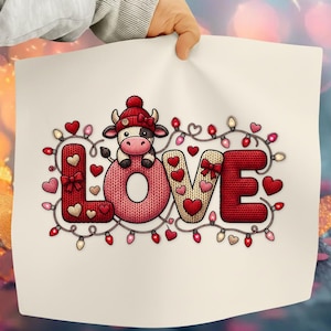 May include: A white print featuring the word "LOVE" with a cow wearing a red hat. The letters are decorated with hearts and surrounded by string lights with red and pink bulbs. The cow is black and white.