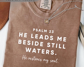 May include: Espresso-colored t-shirt with the text "Psalm 23: He leads me beside still waters. He restores my soul." The shirt has a Comfort Colors tag and a silver necklace. The words "Next Day Shipping" and "Comfort Colors" are also visible.