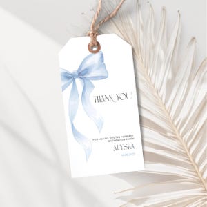 May include: White gift tag featuring a light blue watercolour bow illustration. The tag says "THANK YOU" and includes the text "FOR MAKING THIS THE HAPPIEST BIRTHDAY ON EARTH" and a name and date. A natural twine loop is at the top.