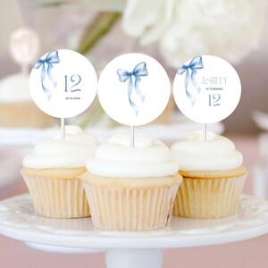 May include: Three cupcakes with white icing are decorated with round toppers. The toppers display a watercolour blue bow and the number 12. One topper reads "Ashley is turning 12". The cupcakes are on a white cake stand.