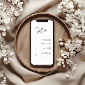 May include: A smartphone displays a wedding invitation with the text "we still do." The invitation includes the names Melissa & Alex, the date September 30, and the venue address. The phone rests on a wooden tray, surrounded by white flowers.
