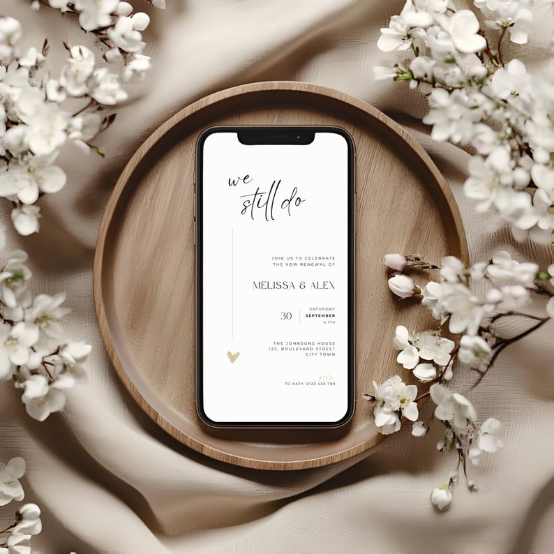May include: A smartphone displays a wedding invitation with the text "we still do." The invitation includes the names Melissa & Alex, the date September 30, and the venue address. The phone rests on a wooden tray, surrounded by white flowers.