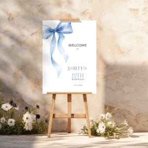 May include: A white sign on a wooden easel featuring a light blue bow illustration. The sign reads "WELCOME TO ASHLEY'S 12TH BIRTHDAY 10.10.2026". The easel is surrounded by white flowers.