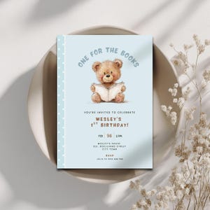 Op de afbeelding: Een lichtblauwe uitnodigingskaart met een aquarel illustratie van een teddybeer die een boek leest. De tekst luidt "ONE FOR THE BOOKS" en "WESLEY'S 1ST BIRTHDAY!" met datum, tijd en RSVP details.
