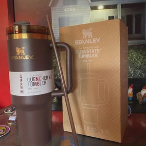 May include: A brown Stanley Flowstate Quencher H2.0 Tumbler with a handle and straw, next to its cardboard box. The box features the Stanley logo and product details. The tumbler is designed for all-day hydration.