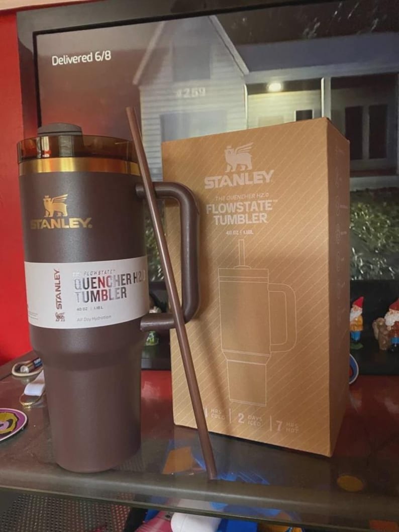 May include: A brown Stanley Flowstate Quencher H2.0 Tumbler with a handle and straw, next to its cardboard box. The box features the Stanley logo and product details. The tumbler is designed for all-day hydration.