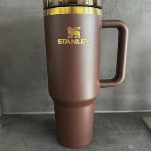 May include: A brown insulated tumbler with a handle and a clear lid. The tumbler has a gold rim and the word "STANLEY" in gold lettering, along with a logo. The tumbler is designed to keep beverages hot or cold.