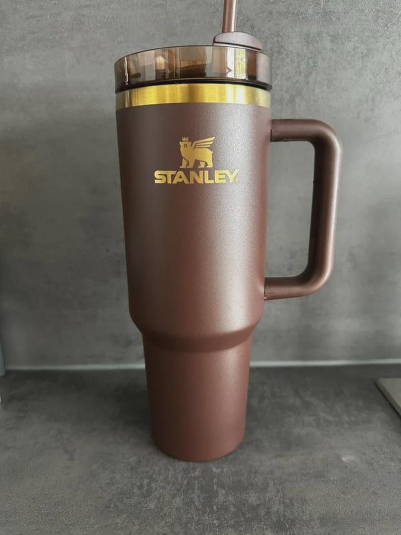 May include: A brown insulated tumbler with a handle and a clear lid. The tumbler has a gold rim and the word "STANLEY" in gold lettering, along with a logo. The tumbler is designed to keep beverages hot or cold.