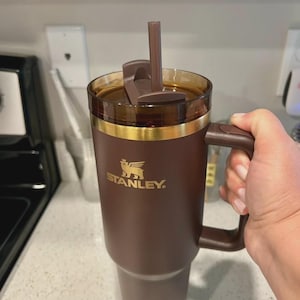 May include: A brown Stanley travel mug with a handle and a straw. The mug has a gold band near the top and the Stanley logo in gold. The lid is brown and has a flip-top closure. The mug is held by a person.