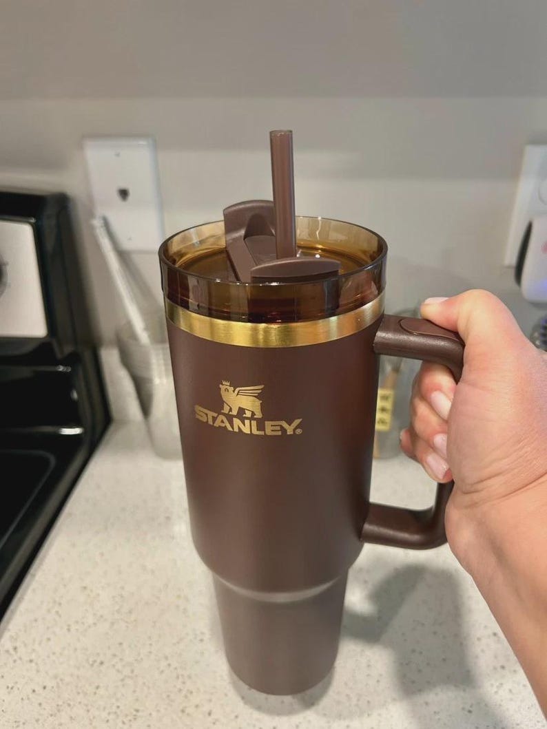 May include: A brown Stanley travel mug with a handle and a straw. The mug has a gold band near the top and the Stanley logo in gold. The lid is brown and has a flip-top closure. The mug is held by a person.