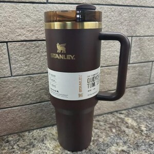 May include: A dark brown Stanley Quencher Tumbler with a handle. The tumbler has a gold rim and a clear plastic lid. The Stanley logo is in gold. The tumbler is 40 oz.