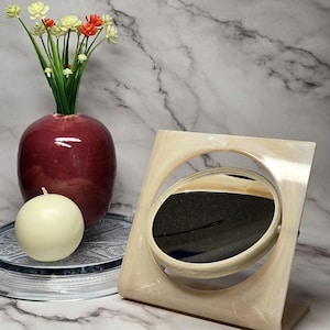 May include: A vanity mirror with a square frame and an oval mirror. The frame has a light, pearlescent finish. A burgundy vase with flowers and a white candle are also in the image, suggesting a decorative theme.
