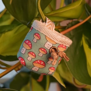Könnte beinhalten: Kleine, quadratische Stofftasche mit einem Muster aus roten und weißen Pilzen auf türkisem Hintergrund. Die Tasche hängt an einem Pflanzenstiel. Die Pflanze hat große, grüne Blätter mit gelben Akzenten.