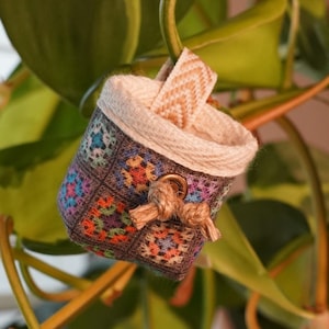 Könnte beinhalten: Ein kleiner, farbenfroher, gehäkelter Korb mit cremefarbenem Rand und einer dekorativen Schleife. Der Korb hängt zwischen den grünen Blättern und Ranken einer Zimmerpflanze. Das Design des Korbes zeigt Quadrate in verschiedenen Farben.