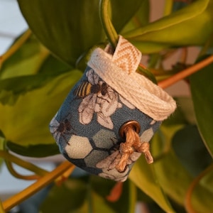 Könnte beinhalten: Kleine blaue Stofftasche mit Bienen- und Wabenmuster, die an einem Pflanzenstiel hängt. Die Tasche hat einen beigen Rand und einen kleinen Holzverschluss. Der Hintergrund zeigt grüne Blätter und Stiele.