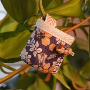 Könnte beinhalten: Ein kleiner, hängender Stoffblumentopf mit einem floralen und wabenförmigen Muster auf marineblauem Hintergrund. Der Topf hängt an einem Pflanzenstiel, umgeben von grünen Blättern. Er hat eine weiße Bordüre und ein kleines Seildetail.