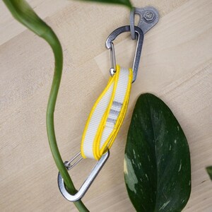Könnte beinhalten: Ein silberner Karabiner mit einem gelb-weiß gestreiften Band, das an einem silbernen Haken befestigt ist. Der Karabiner hängt an einer Wand.