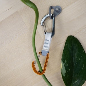 Könnte beinhalten: Ein grauer und orangefarbener Karabiner mit einem weißen Band, das an einer grauen Wandhalterung befestigt ist. Der Karabiner hängt an einer grünen Pflanze.