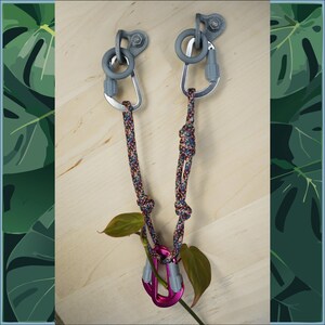 Könnte beinhalten: Zwei graue Metallhaken mit einer Schlaufe und einem Karabiner, die an jedem befestigt sind. Die Karabiner sind durch ein mehrfarbiges Seil verbunden. Ein rosa Karabiner ist an dem Seil befestigt.