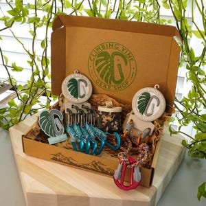 Könnte beinhalten: Ein brauner Karton mit dem Logo "Climbing Vine Gear Shop" auf der Oberseite. Die Box ist geöffnet und enthält verschiedene Kletterausrüstung, darunter Karabiner, Seil und eine kleine Tasche. Die Box steht auf einer Holzoberfläche.