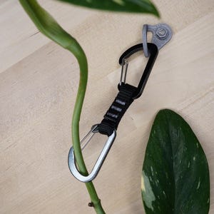 Könnte beinhalten: Ein schwarzer und silberner Pflanzenhänger mit einem Karabinerhaken, der an einem grünen Pflanzenstängel befestigt ist. Der Hänger ist mit einem silbernen Haken an einer Wand befestigt.