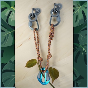 Könnte beinhalten: Zwei graue Metall-Kletterkarabiner mit einem blauen Karabiner, der an einem mehrfarbigen Seil befestigt ist. Das Seil ist an zwei grauen Metallhaken befestigt, die an einer Holzoberfläche montiert sind.