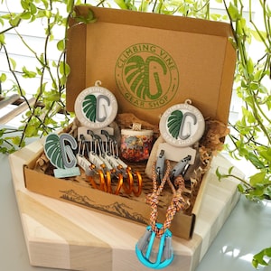 Könnte beinhalten: Ein brauner Karton mit dem Text "Climbing Vine Gear Shop" und einem grünen Monstera-Blatt-Logo. Die Box ist geöffnet und enthält verschiedene Kletterausrüstung, darunter Karabiner, Schlaufen und eine kleine Tasche mit Blumenmuster.