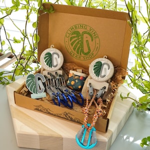 Könnte beinhalten: Ein brauner Karton mit der Aufschrift "Climbing Vine Gear Shop" auf der Oberseite. Die Box ist geöffnet und enthält verschiedene Kletterausrüstung, darunter Karabiner, Schlaufen und eine kleine Tasche mit Weltraumdesign.