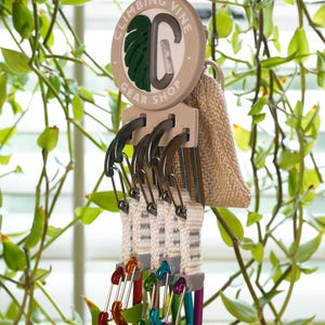 Könnte beinhalten: Ein Kletterausrüstungsorganisator mit dem Text "CLIMBING VINE GEAR SHOP" auf einem kreisförmigen Schild. Der Organizer enthält mehrere Karabiner in verschiedenen Farben, darunter Rot, Orange, Grün, Blau und Lila. Ein kleiner Jutesack hängt an der Seite.