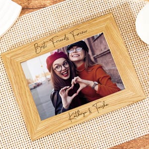 May include: A wooden photo frame with the text "Best Friends Forever" and the names "Kathryn & Taisha" engraved on the bottom. The frame holds a photo of two friends smiling and making a heart shape with their hands.