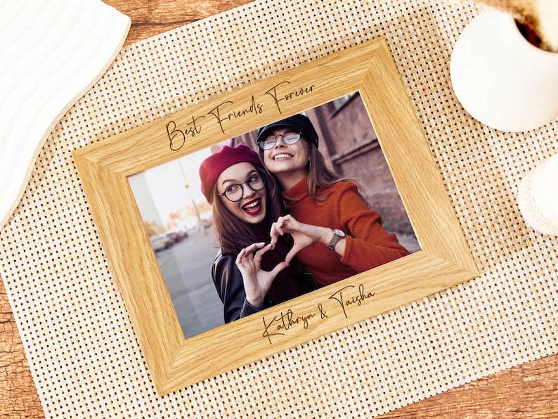 May include: A wooden photo frame with the text "Best Friends Forever" and the names "Kathryn & Taisha" engraved on the bottom. The frame holds a photo of two friends smiling and making a heart shape with their hands.
