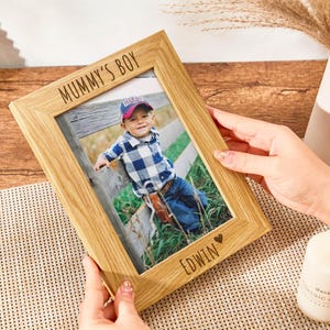 May include: A wooden photo frame with a light brown finish. The frame has a white inscription that reads "Mummy's Boy" and a smaller inscription that reads "EDWIN" with a heart symbol. The frame contains a photo of a young boy wearing a plaid shirt and jeans.